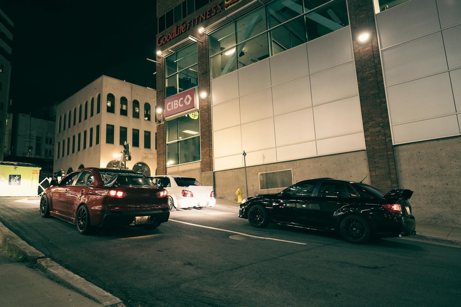 A nighttime scene of parked cars on an urban street outside a fitness center and bank.