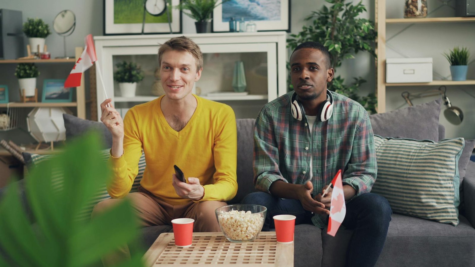 Two friends sitting on a sofa, enjoying snacks, holding Canadian flags indoors.